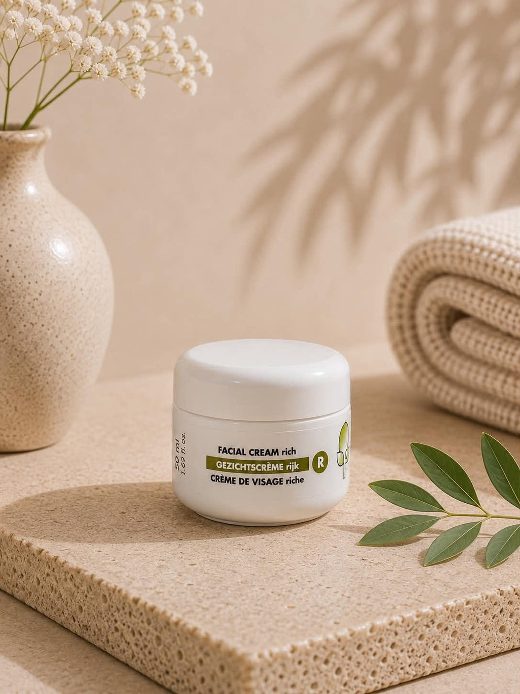 Pot de crème visage riche Jojoba Care sur un socle naturel avec fleurs et serviette.