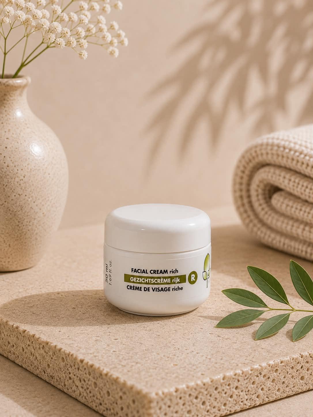 Pot de crème visage riche Jojoba Care sur un socle naturel avec fleurs et serviette.